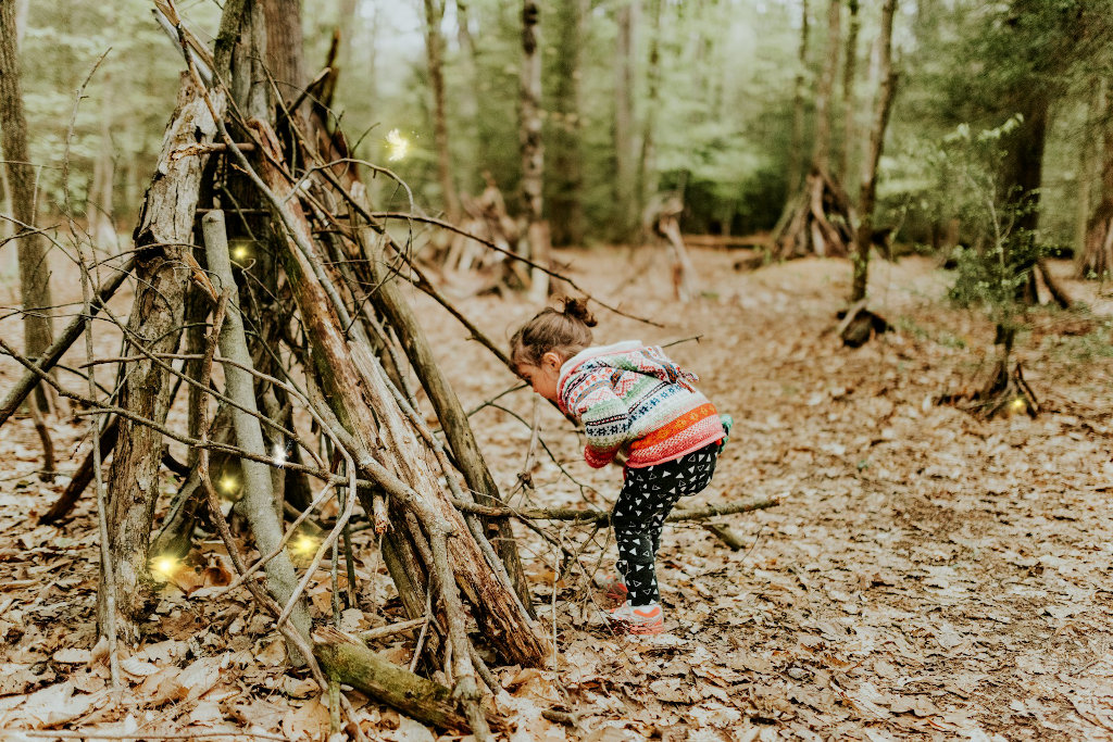 little girl looking curious into a structure made of branches in a forest